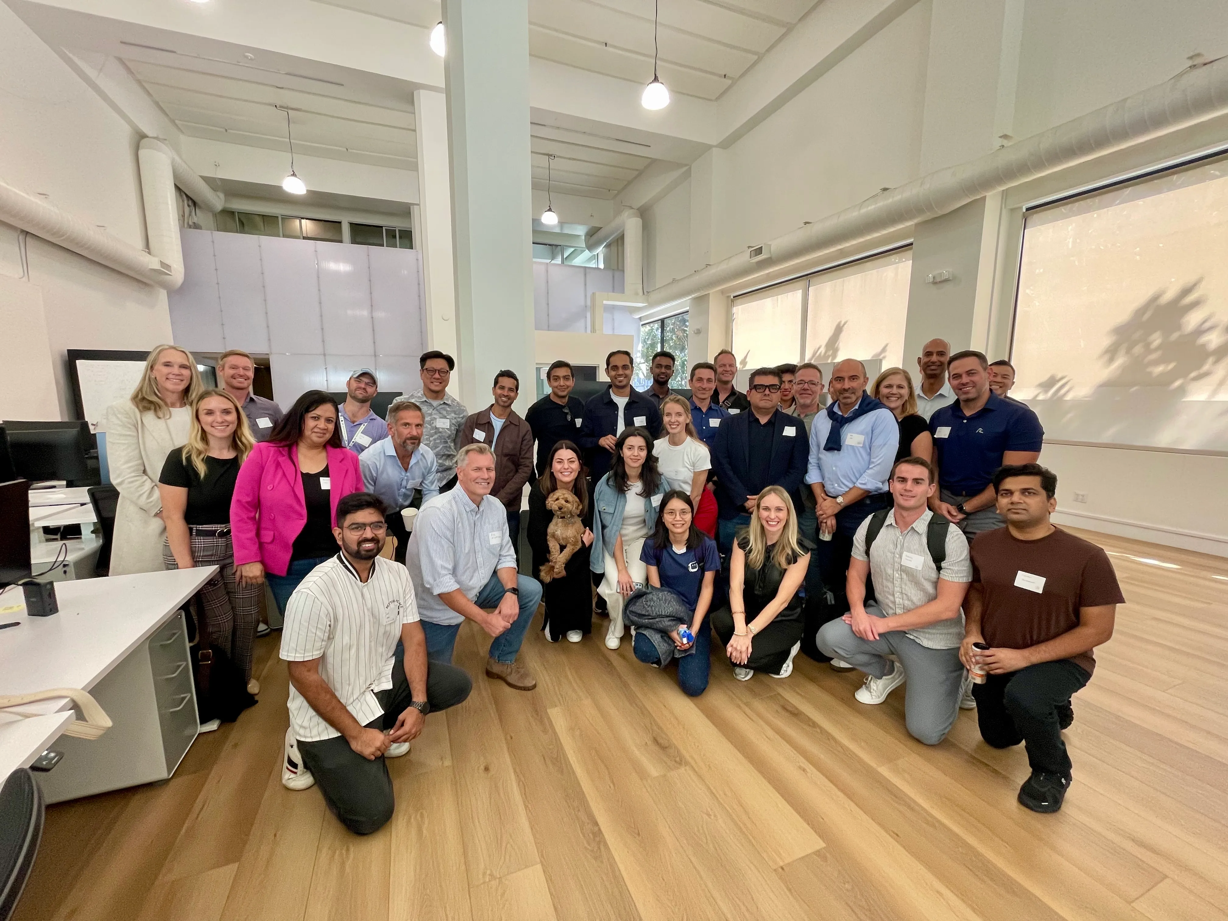 A large group of founders and operators inside a sunlit SF workspace, with a small dog in the front row